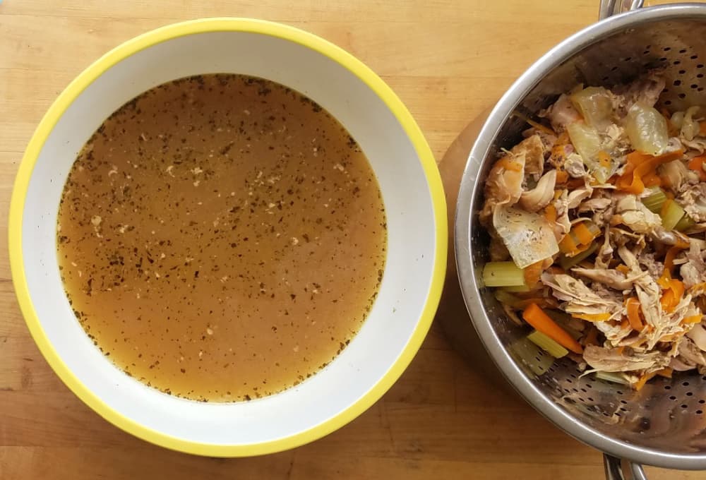 Strained chicken broth in large bowl, with colander to the side, filled. 