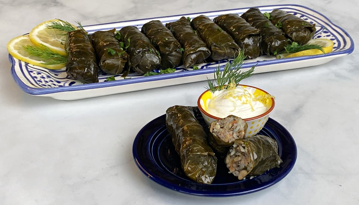 Serving of dolma on small plate, with a small cup of yogurt on the side. Tray of rolled grape leaves in the background.