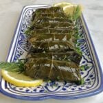 Close-up of rolled, stuffed grape leaves lined up a long serving platter. Garnished with lemon slices and fresh herbs.
