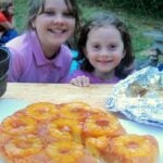 Two girls looking across a wooden table to pineapple-upside-down cake in foreground.
