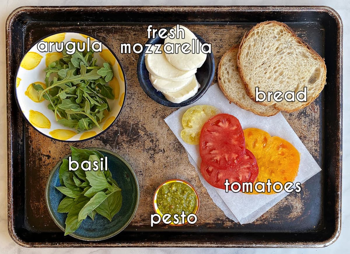Caprese sandwich ingredients, labeled: bread, tomatoes, mozzarella, basil, arugula, and pesto.