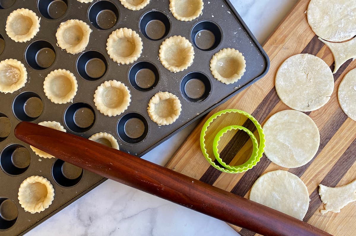 corner of a large mini-muffin tin with pastry formed in every other hole. Rolling pin and rolled pastry on a cutting board to the side.