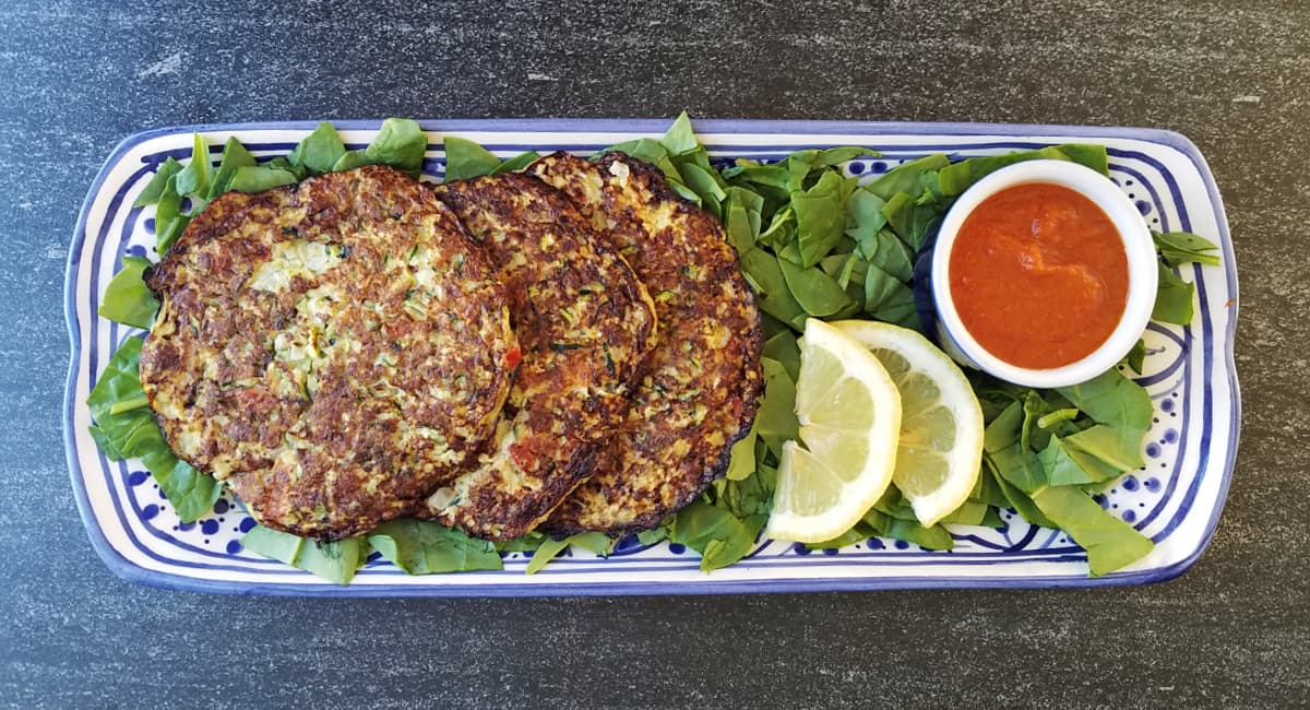 Overhead shot zucchini cakes with sauce, lemon slices, and a small ramakin of sauce on a rectangular serving tray. 