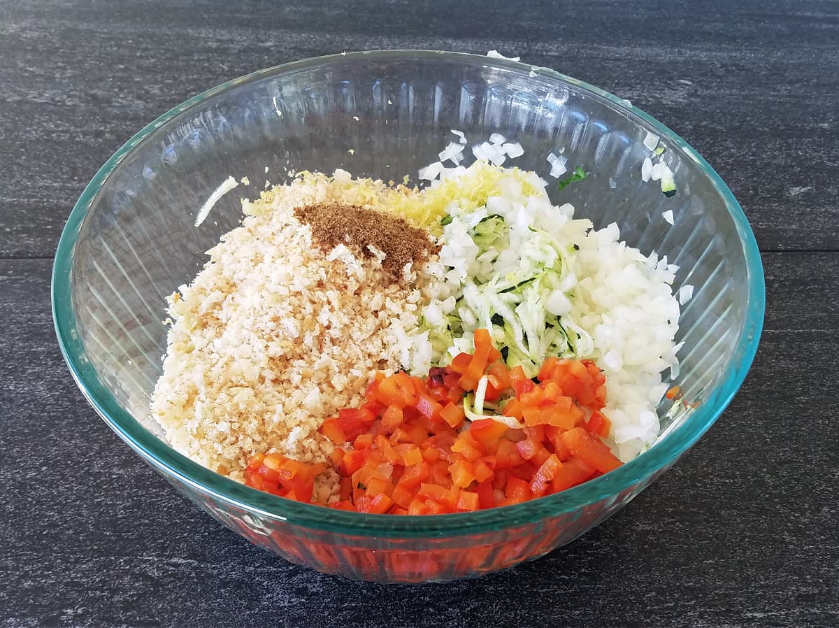 Glass mixing bowl with ingredients: chopped red pepper, squeezed zucchini, onion, Panko crumbs, parmesan, salt and pepper. 