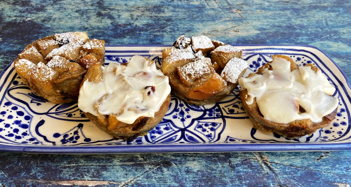 Four bread pudding muffins aligned horizontally on a small, rectangular serving tray. Two are frosted, and two are dusted with powdered sugar.