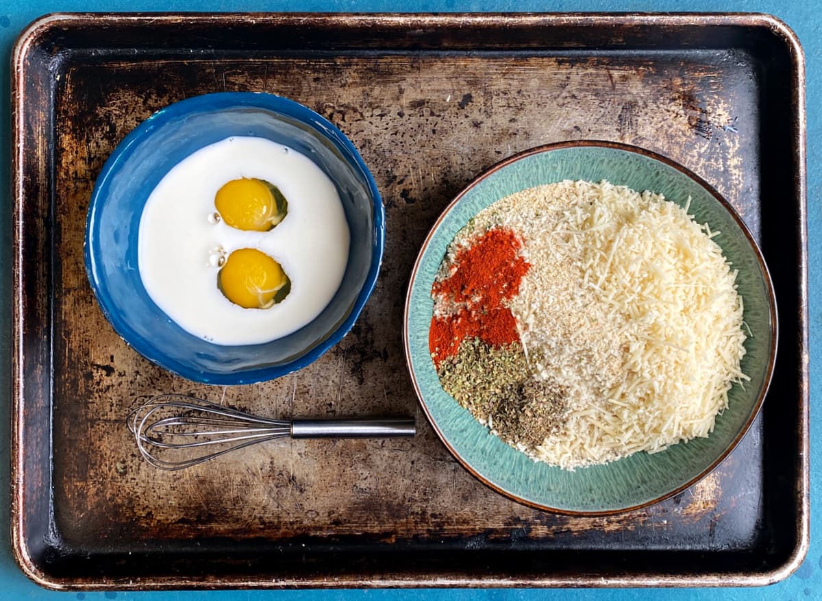 Two bowls on a baking tray: one with eggs and milk, and the other with bread crumbs and herbs.