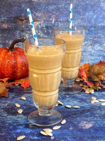 Two pumpkin smoothies in old-fashioned soda glasses. Pumpkin seeds sprinkled around the foot of glasses. Fall decor (leaves, pumpkin) in background, and polka dot straw are in the glasses..