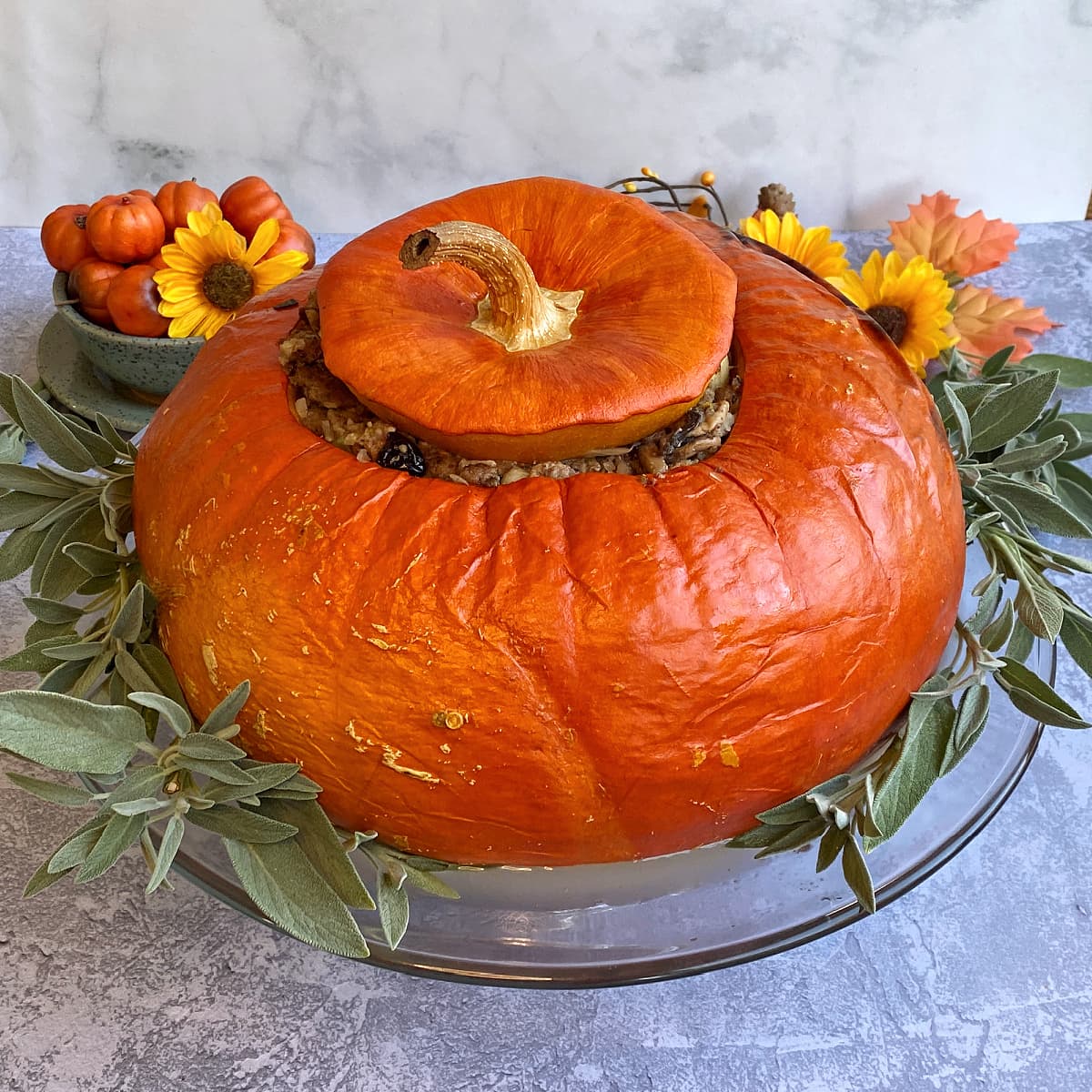 Whole stuffed pumpkin on glass stand, surrounded by fresh sage. 