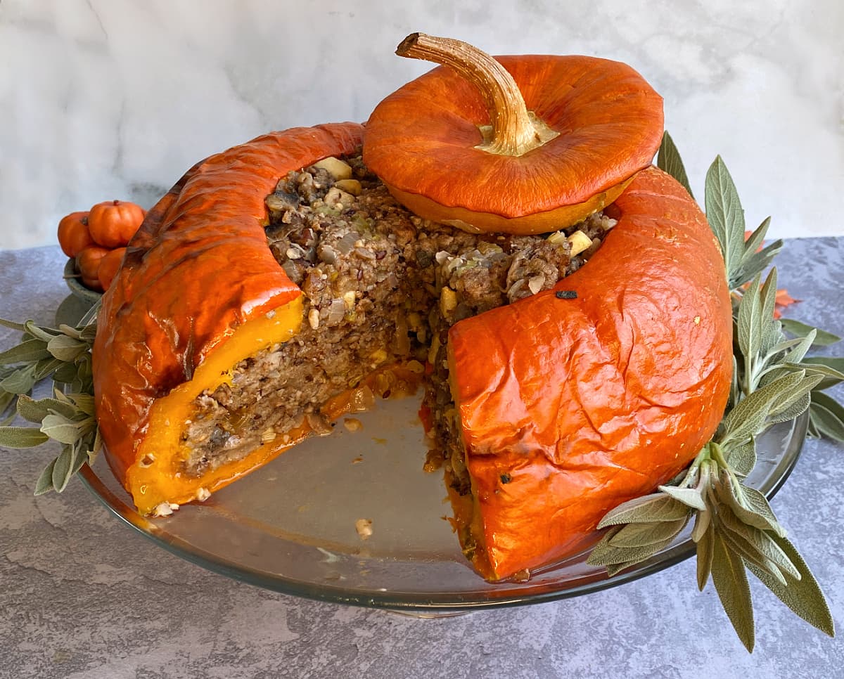 Baked stuffed pumpkin on a glass stand, surrounded by fresh sage, with a large slice cut up.