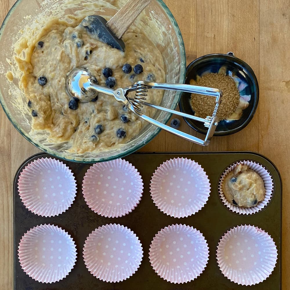 Overhead of muffin cups in tin, and bowl of batter with cookie scoop, ready to fill cups. 