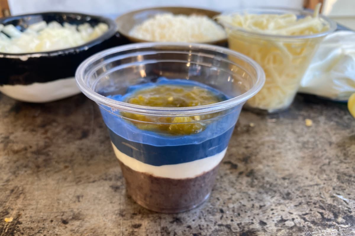 Partially made 7-layer dip in a plastic cup, with salsa in center recess.