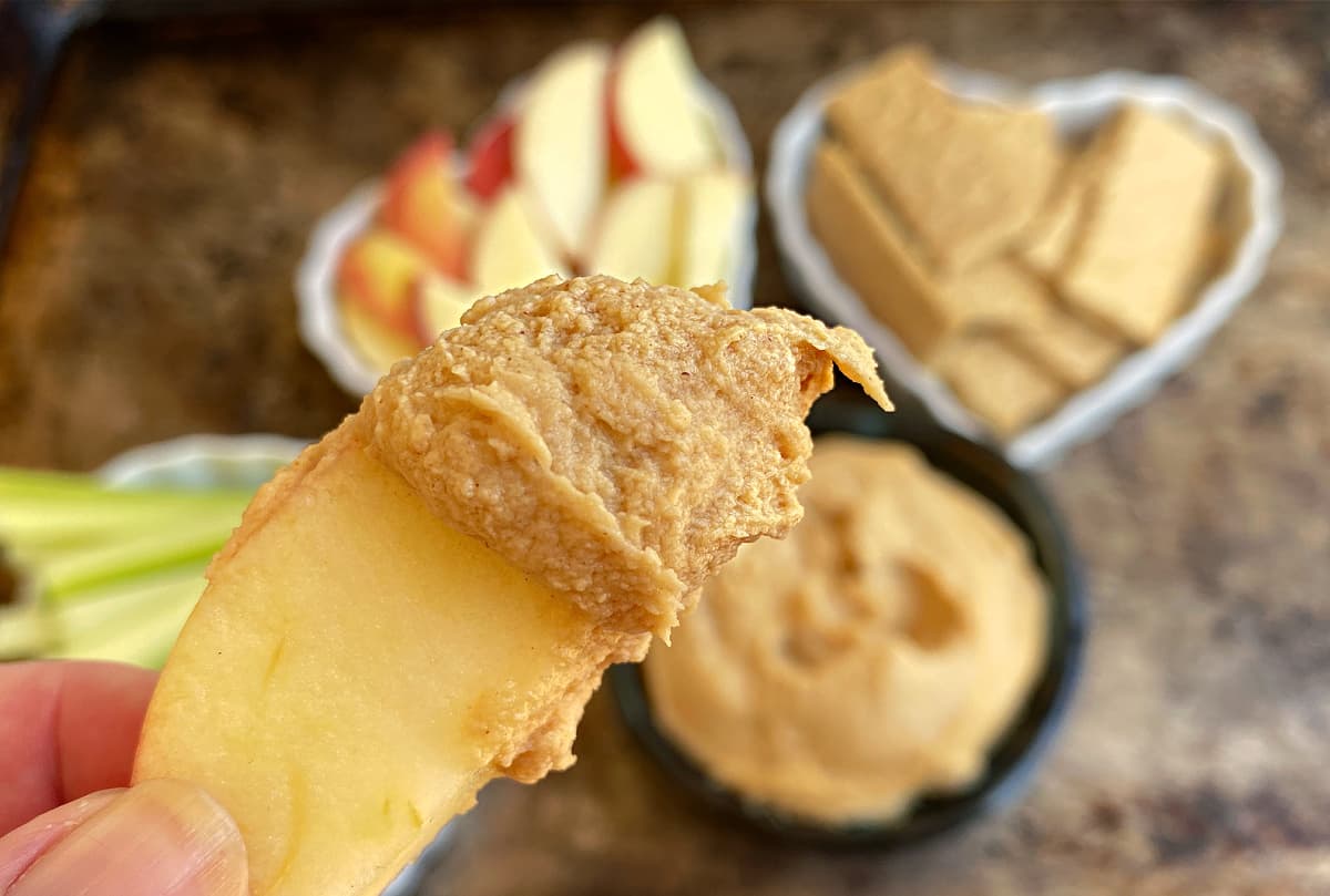 Apple slice dipped in peanut butter dip. Small bowls of apples, graham crackers, and celery in background.