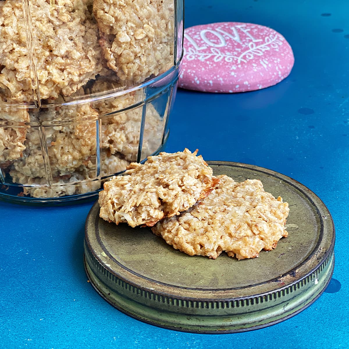 Coconut Oatmeal Cookies in glass jar. Two stacked on lid that is setting on the counter. 