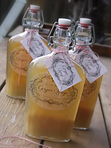 Three old-time stopper bottles filled and labeled "Ginger Syrup" written on paper tags, and tied on with red-striped bakers twine.