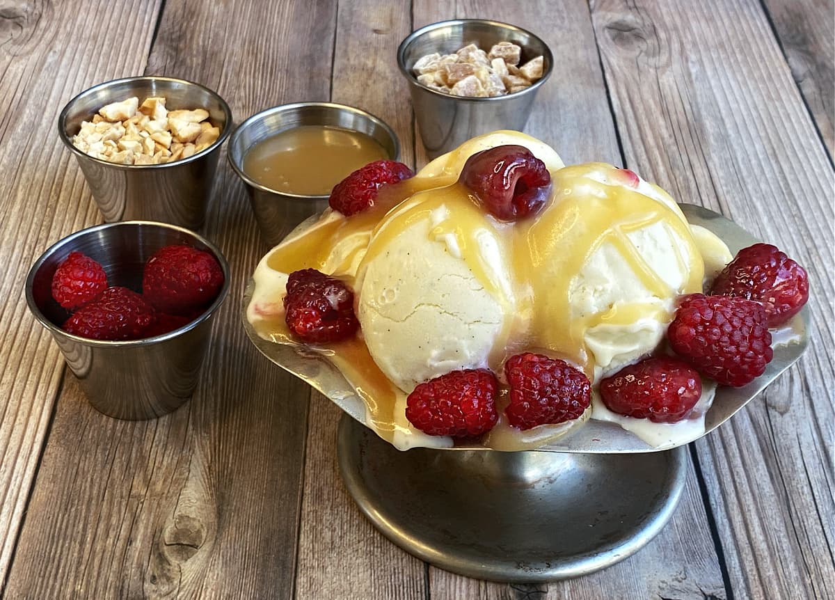 Old-fashion ice cream sundae dish filled with vanilla ice crea, and topped with caramel and raspberries. Service cups of additional toppings in background.