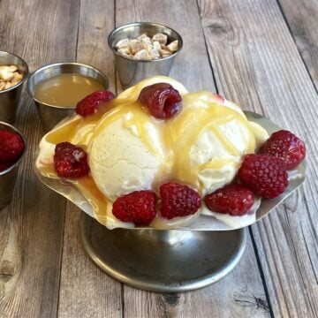 Old-fashion ice cream sundae dish filled with vanilla ice cream, and topped with caramel and raspberries. Service cups of additional toppings in background.