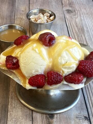 Old-fashion ice cream sundae dish filled with vanilla ice cream, and topped with caramel and raspberries. Service cups of additional toppings in background.