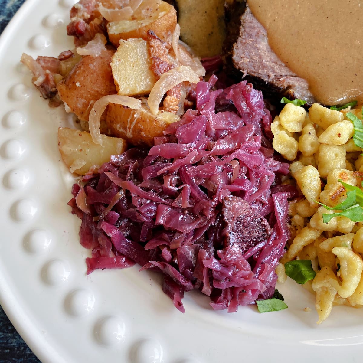 Geram red cabbage centered on a plate with German potato salad, sauerbraten, and spaetzle. 