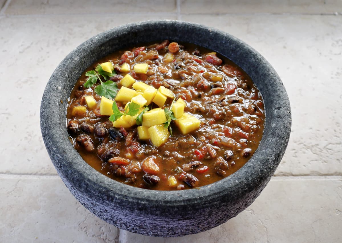 Vegan chili with beans in a stone bowl, garnished with diced mango and cilantro.