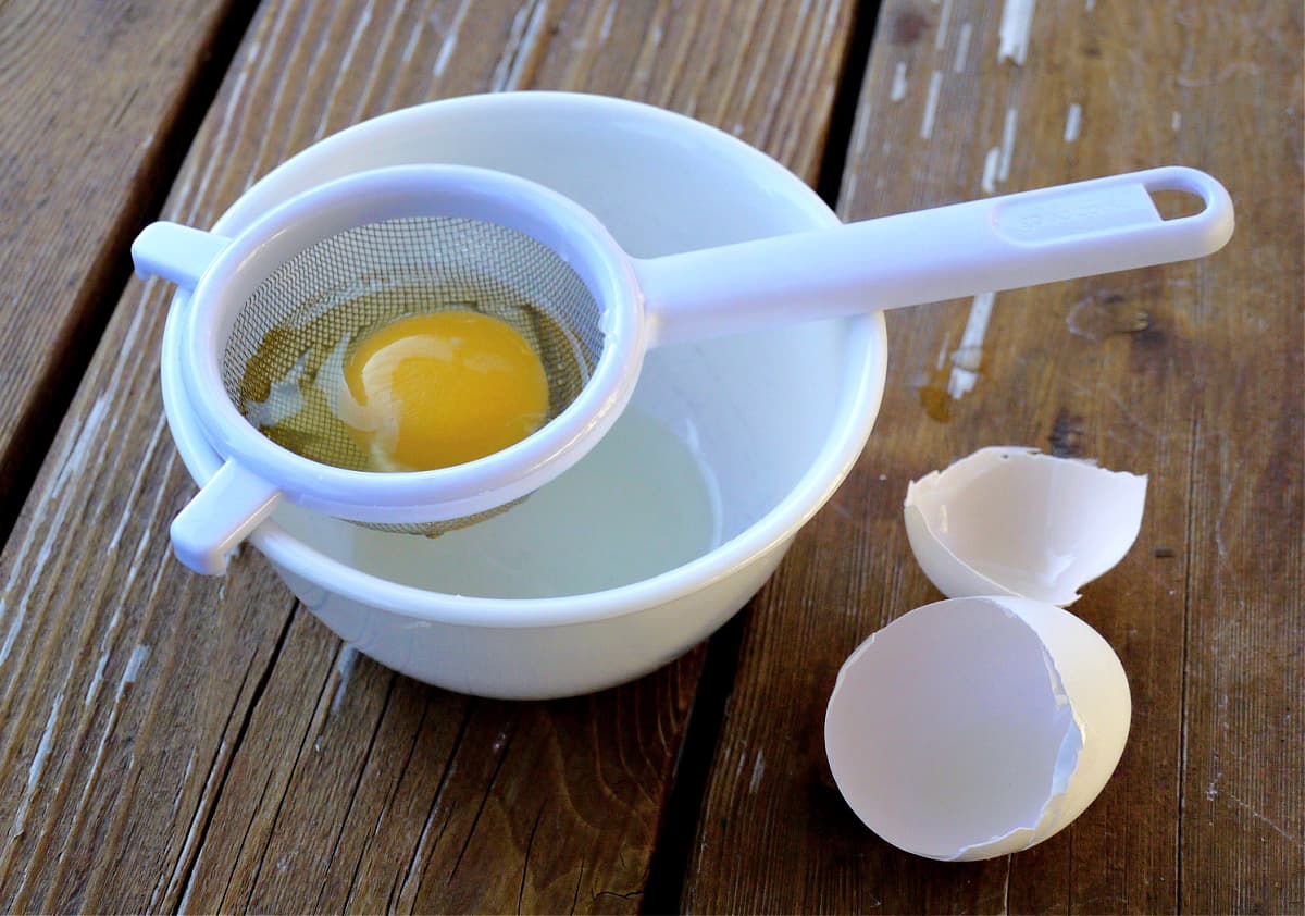 An egg being strained in a small wire strainer over a bowl. 