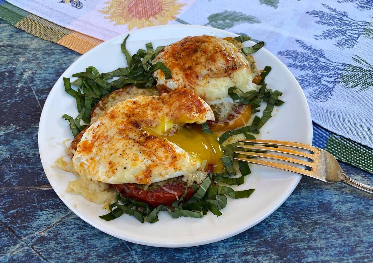 Eggs provencal on a plate, garnished with fresh basil, egg yolk dripping out after being cut with fork.