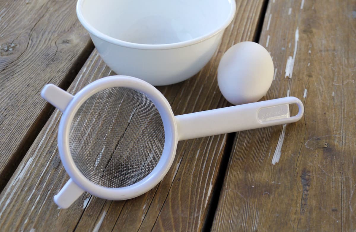 Small strainer with handle beside a small white bowl and a single egg. 
