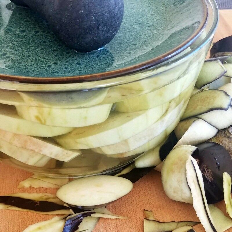Eggplant slices soaking in bowl, with larger plate weighting it down. 