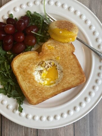 Grilled cheese sandwich with a hole cut out of the middle and an egg cooked it in. Yolk is runny, and the sandwich hole sets to the side with yolk on it. Grapes and arugula on the side of the plate.