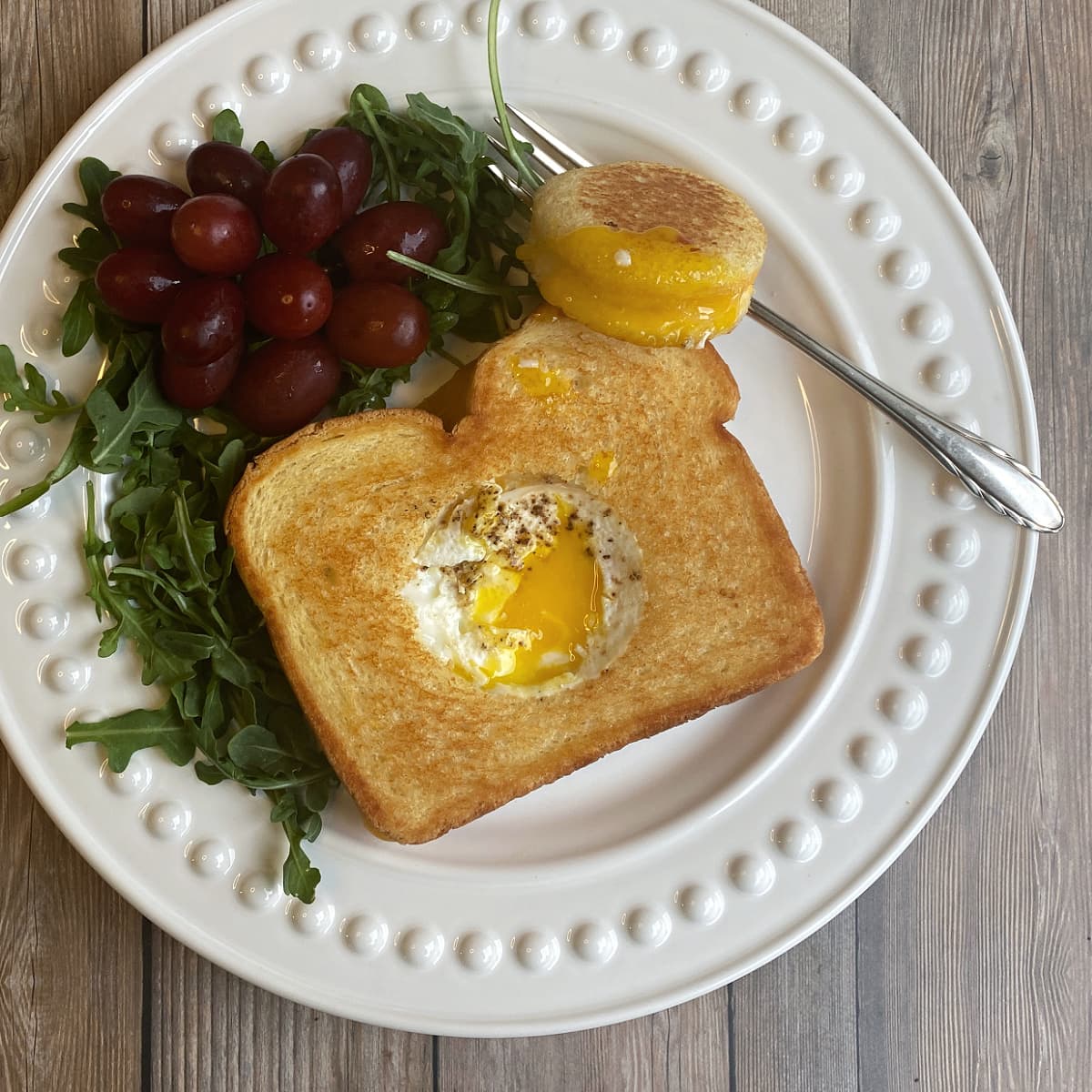 Grilled cheese sandwich with a hole cut out of the middle and an egg cooked it in. Yolk is runny, and the sandwich hole sets to the side with yolk on it. Grapes and arugula on the side of the plate. 