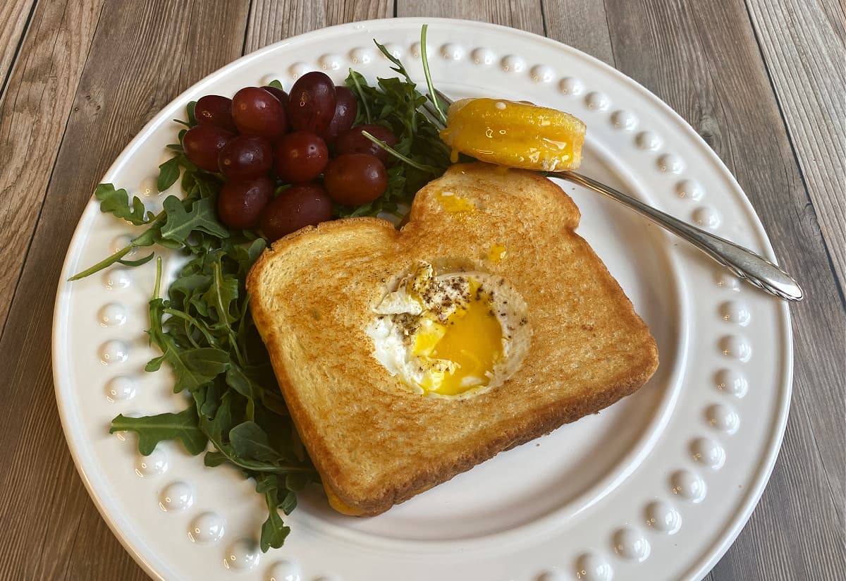 Grilled cheese sandwich with a hole cut out of the middle and an egg cooked it in. Yolk is runny, and the sandwich hole sets to the side with yolk on it.