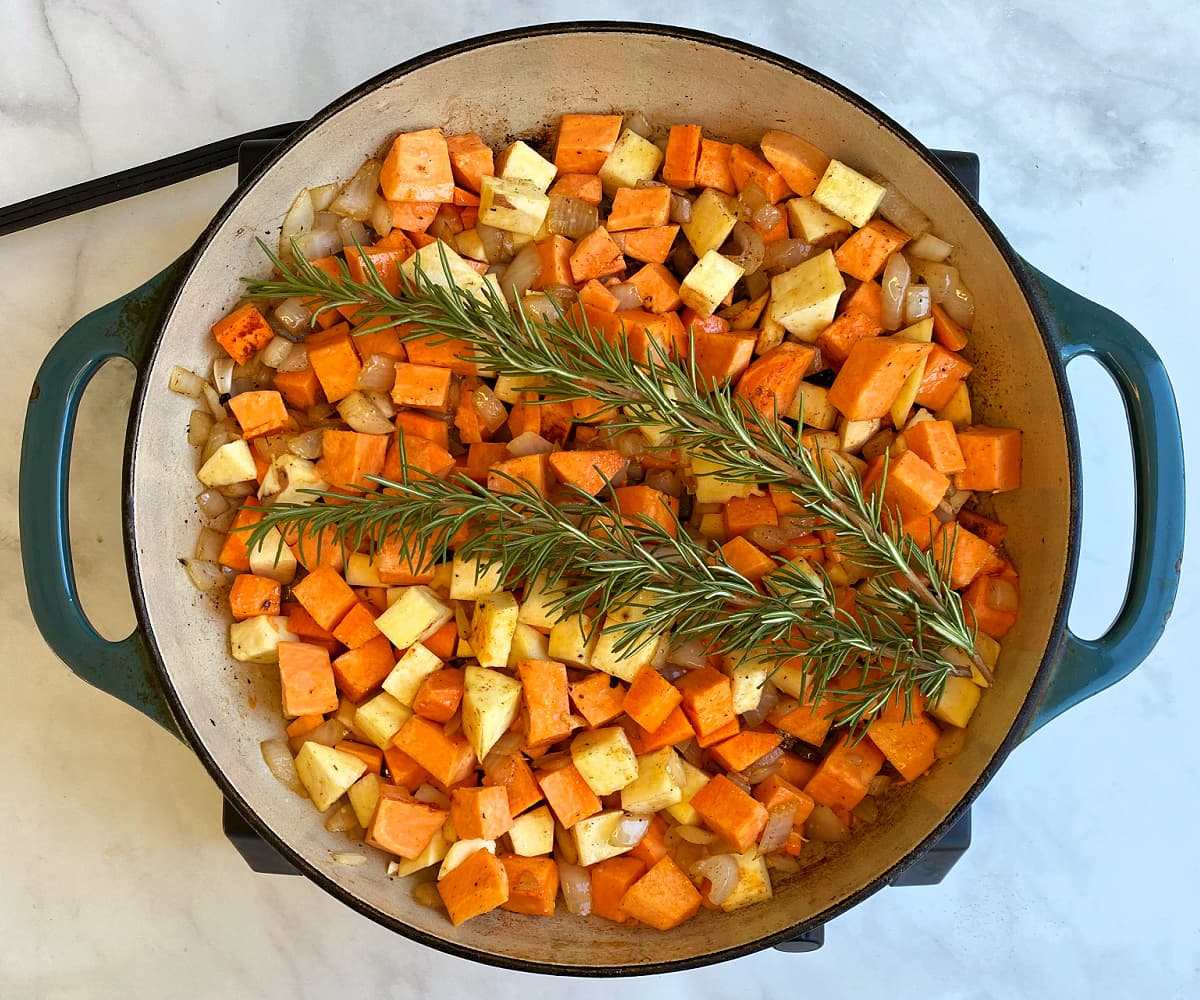 Two sprigs of rosemary laid across the top of cooked hash. Rosemary is fresh and vegetables appear uncooked.