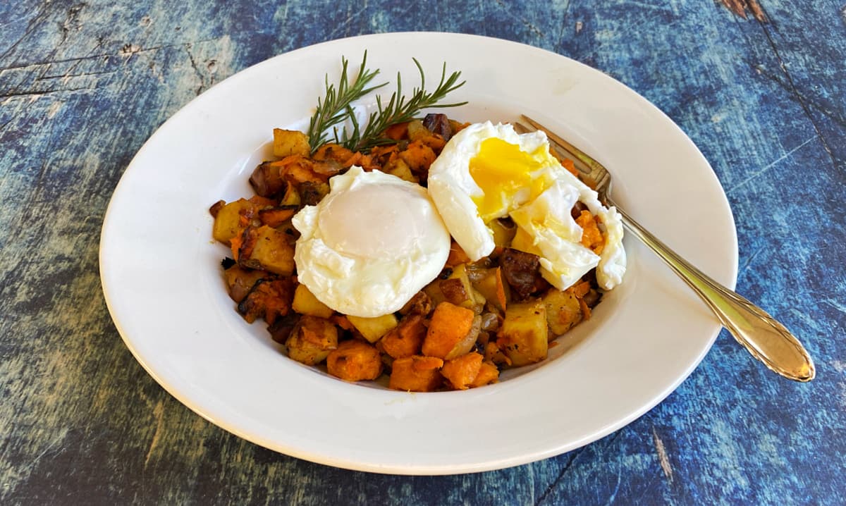 Bowl of sweet potato hash with two poached eggs on top. One egg yolk is broken.
