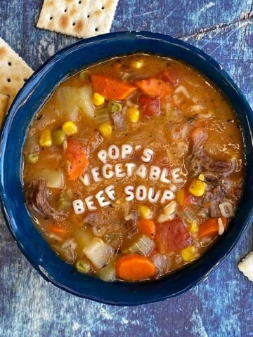 A bowl of vegetable soup, with saltine crackers strewn at the foot of the bowl. Alphabet letters spell out "Pop's Vegetable Beef Soup" across the top of the soup.