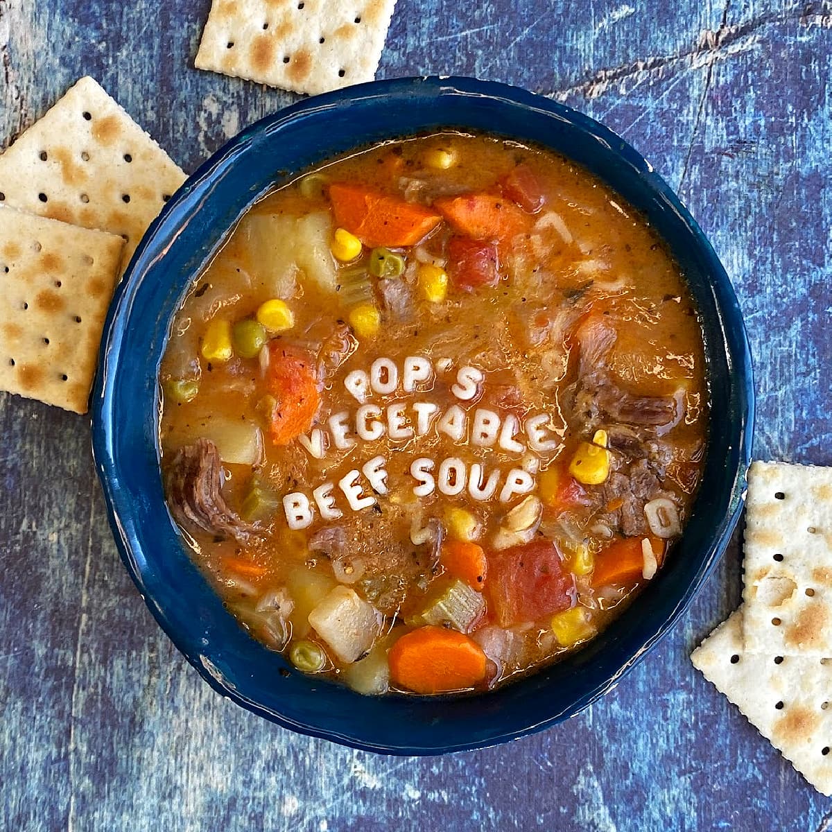 A bowl of vegetable soup, with saltine crackers strewn at the foot of the bowl. Alphabet letters spell out "Pop's Vegetable Beef Soup" across the top of the soup. 