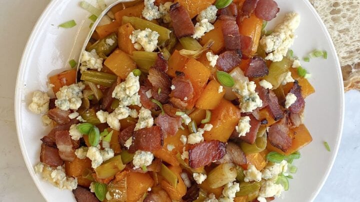 Overhead shot of a plate of prepared butternut squash with leeks, bacon, and blue cheese. Slices of sourdough bread to the side of the plate, and a serving spoon in the mix.