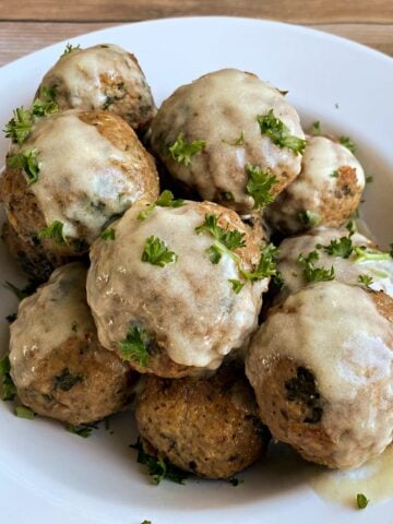 Many meatballs in a bowl, with a sauce poured over and garnished with fresh parsley.