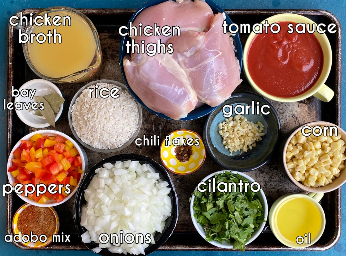 Arroz con Pollo Ingredients on baking tray: chicken thighs, oil, garlic, peppers, corn, onion, rice, chicken broth, tomato sauce, chili flakes, bay leaves, fresh cilantro.