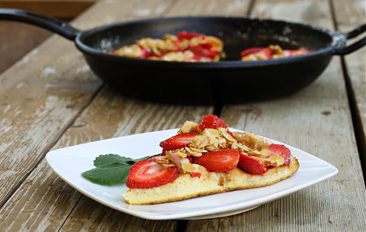 Single slice of strawberry covered omelet on a white plate. Cast iron skillet with remaining omelet in the background.