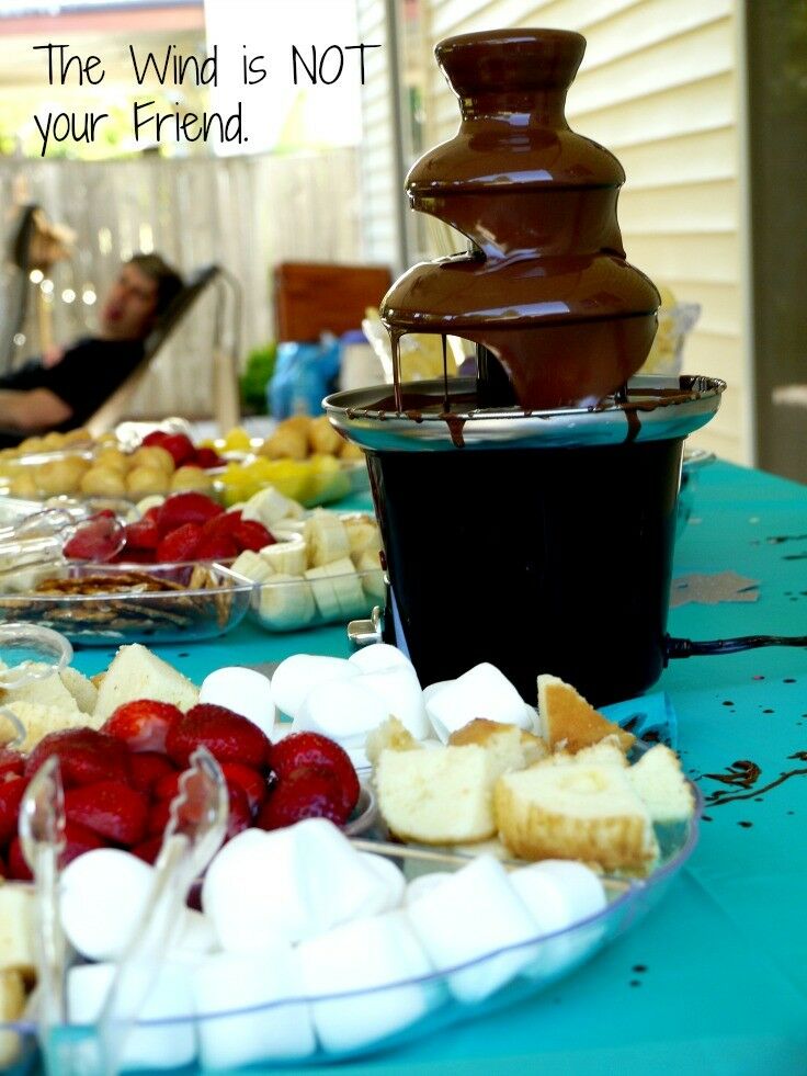 Table of plates filled with fruits, marshmallows and other dippers. Chocolate fountain chocolate splatters on the table. Text overlay reads: The Wind is NOT your friend!