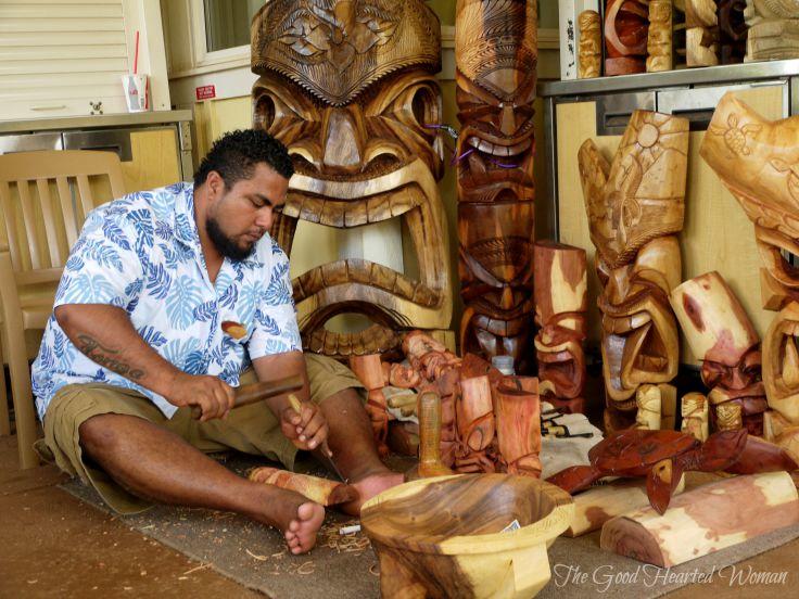 Bearded man wearing Hawaiian shirt, sitting on ground and carving.