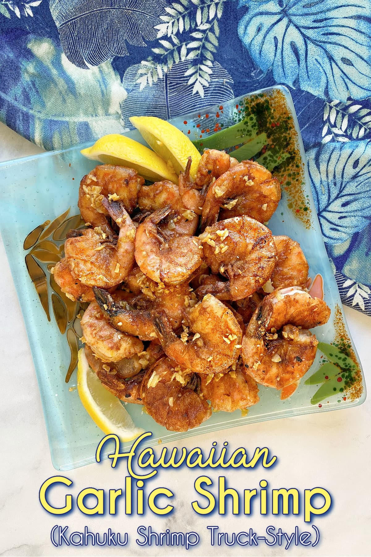 Plate of garlic shrimp, garnished with lemon slices. Hawaiian fabric in background. Pin text reads: Hawaiian Garlic Shrimp (Kahuku Shrimp Truck-style)