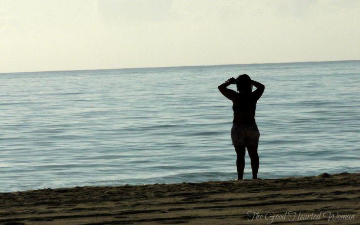 Woman in silhouette looking out at the ocean, both hands up, elbows out, shading her face. 