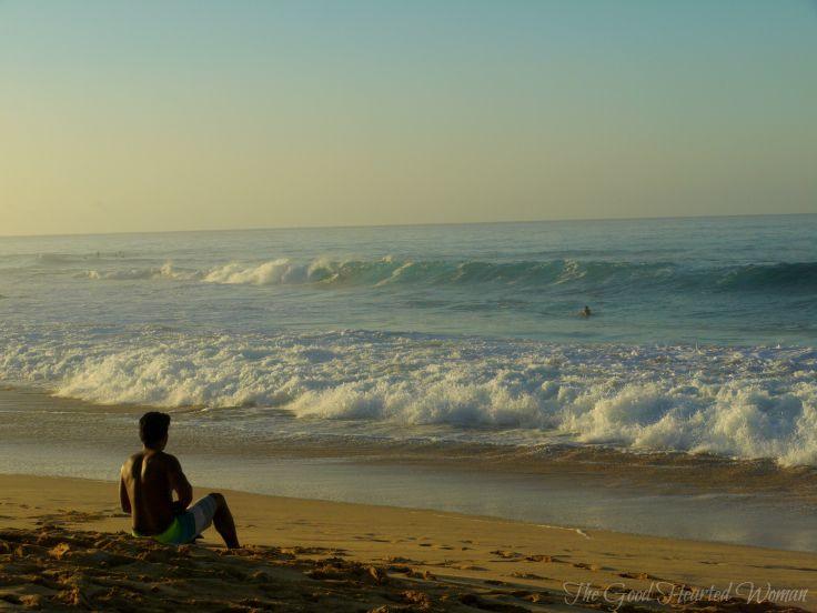 Man sitting on the beach, looking out at the ocean waves. 