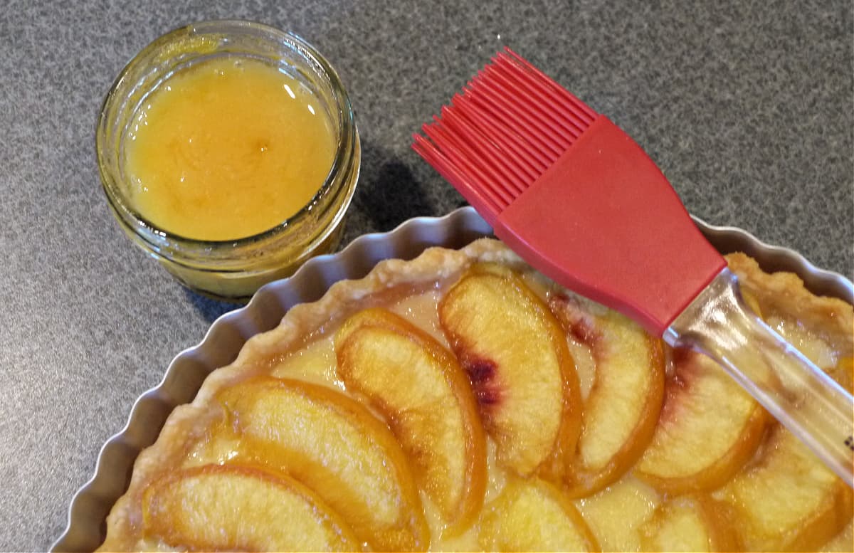 Corner of unbaked peach tart, with a jar of peach jam in the corner, and a red silicone pastry brush resting on edge.