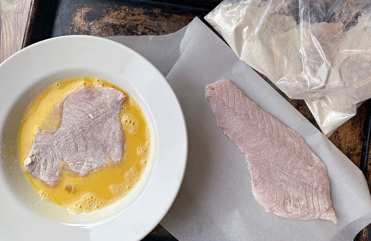 3 raw fish fillets: raw, flour dredged in plastic bag, and egg-dipped in large white bowl.