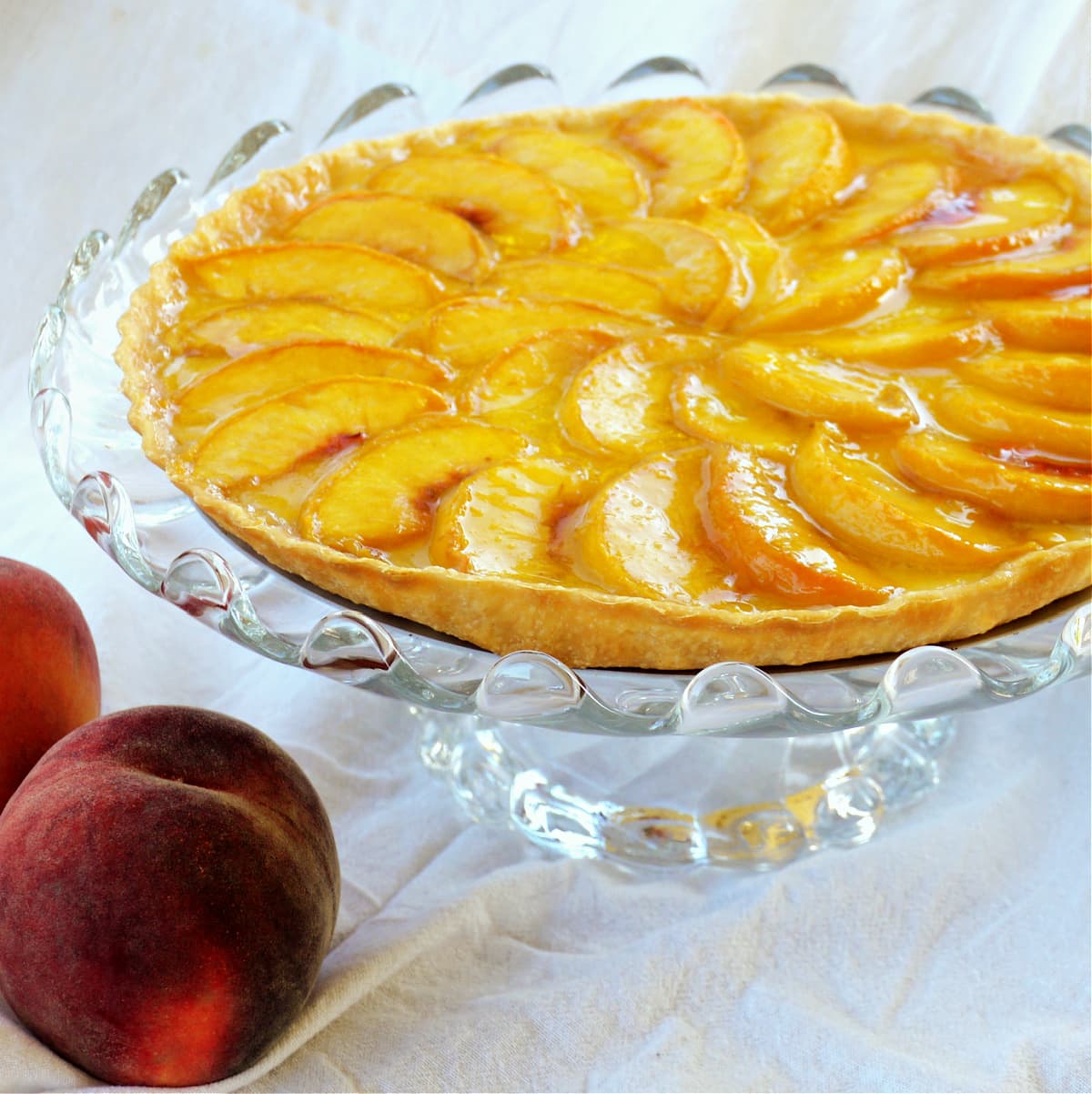 Close-up of a fresh peach tart baked and resting on a glass cake stand. Two whole, ripe peaches in foreground.