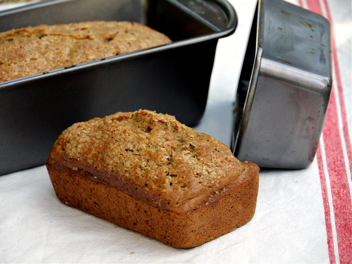 Small loaf of quick bread, unsliced. Empty pan to the side. Larger loaf still in the pan behind the small loaf.