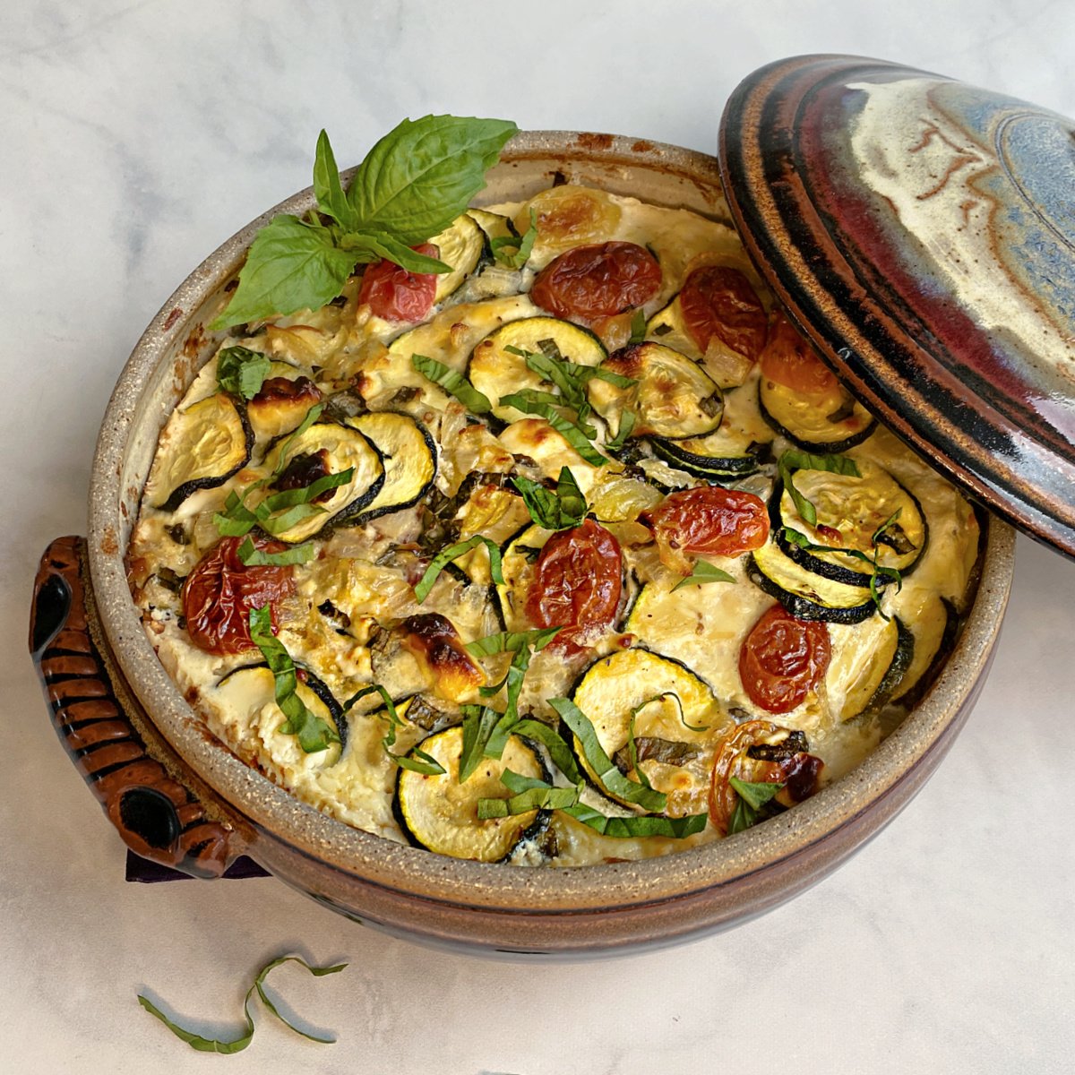 Baked zucchini casserole with tomatoes in a crockery casserole dish, garnished with chiffonaded basil. Casserole lid leaning on the side.