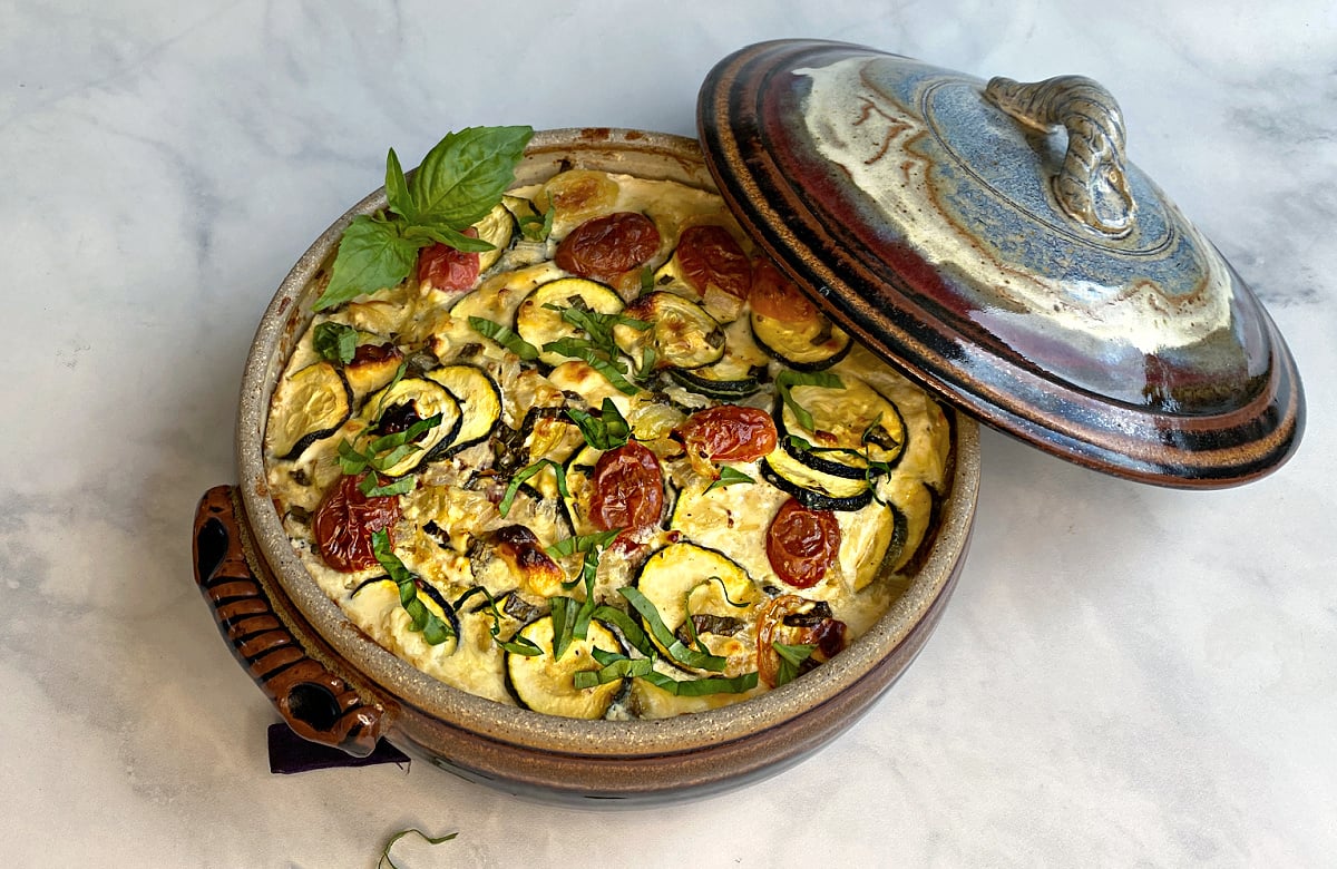 Baked zucchini casserole with tomatoes in a crockery casserole dish, garnished with chiffonaded basil. Casserole lid leaning on the side.