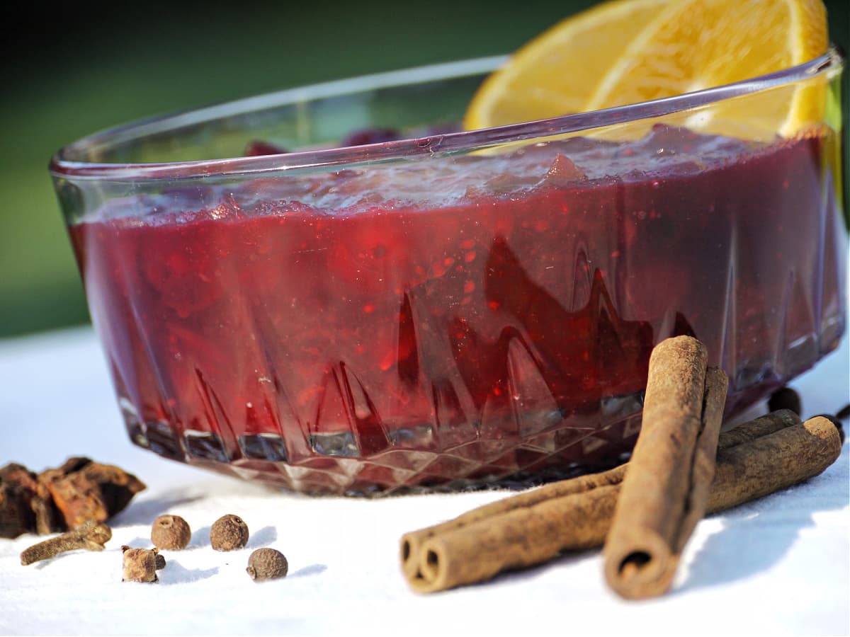 Close-up of cranberry sauce in glass dish, surrounded by whole cinnamon sticks, allspice, cloves, and star anise. 